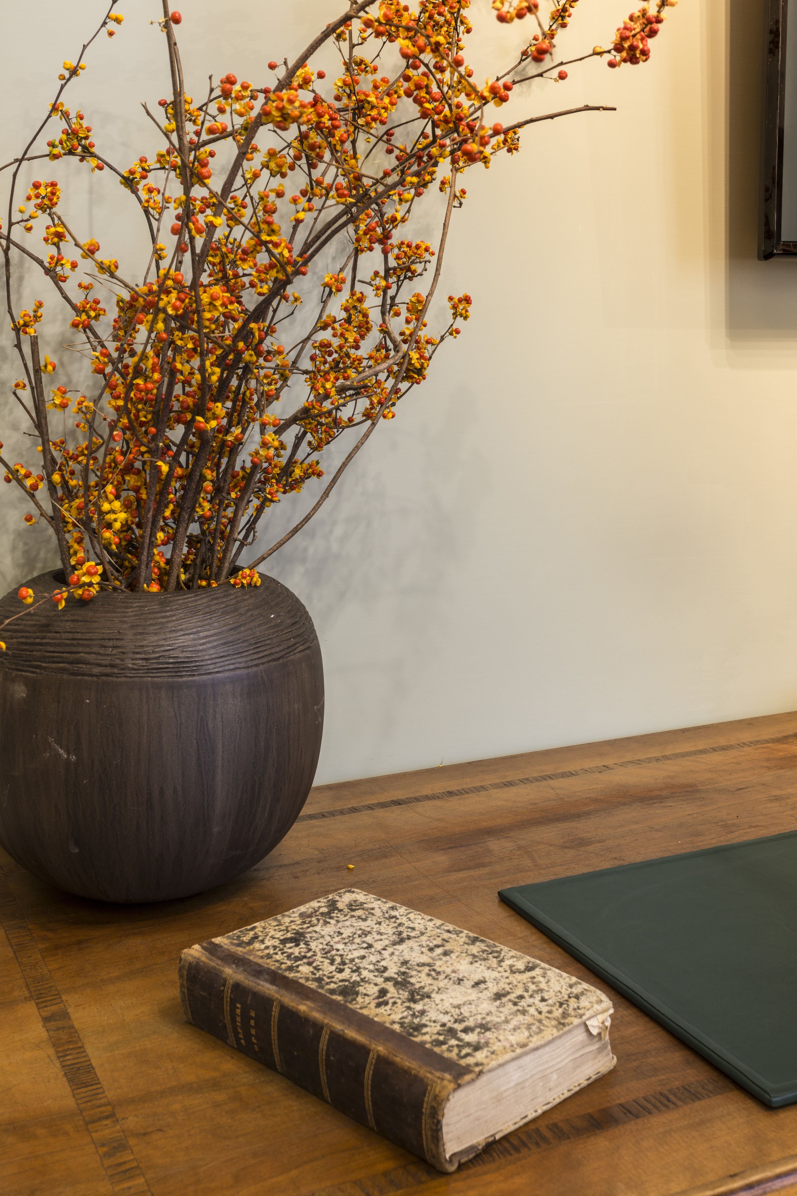 A large vase with branches of yellow and orange flowers, a book, and a green book on a wooden table.