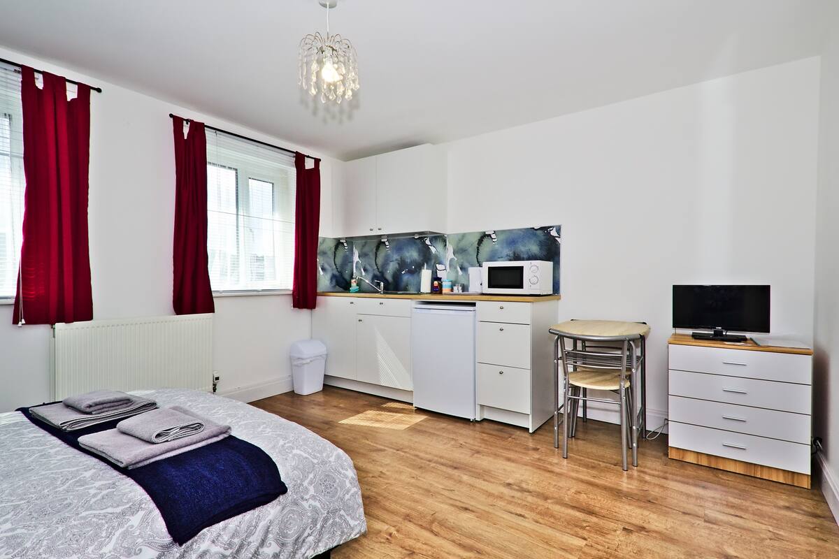A modern bedroom with a bed, a small kitchenette, a TV, and a window with red curtains.