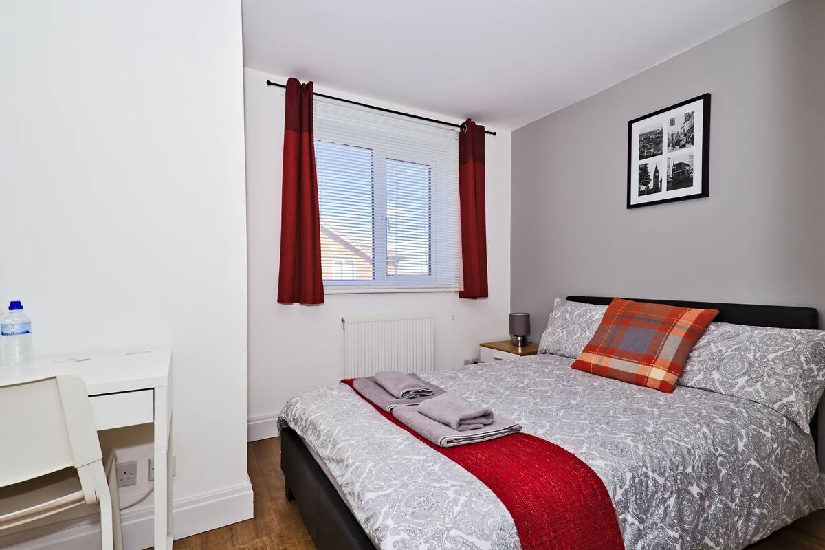 A neatly arranged bedroom with a bed, a bedside table, a desk, and a window with curtains.
