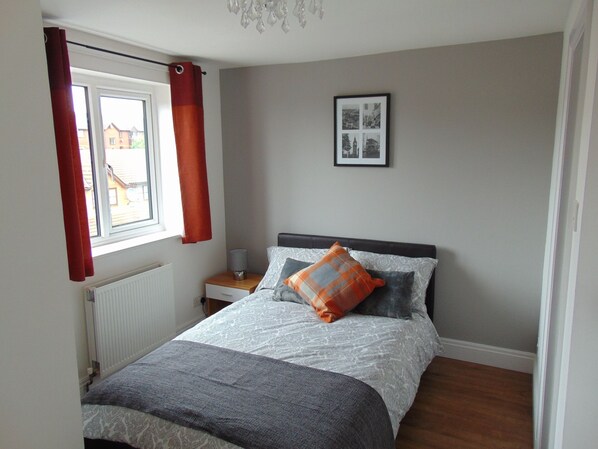 A bedroom with a bed, a nightstand, a radiator, a window with curtains, and a framed picture on the wall. - Harlinger Lodge (London)