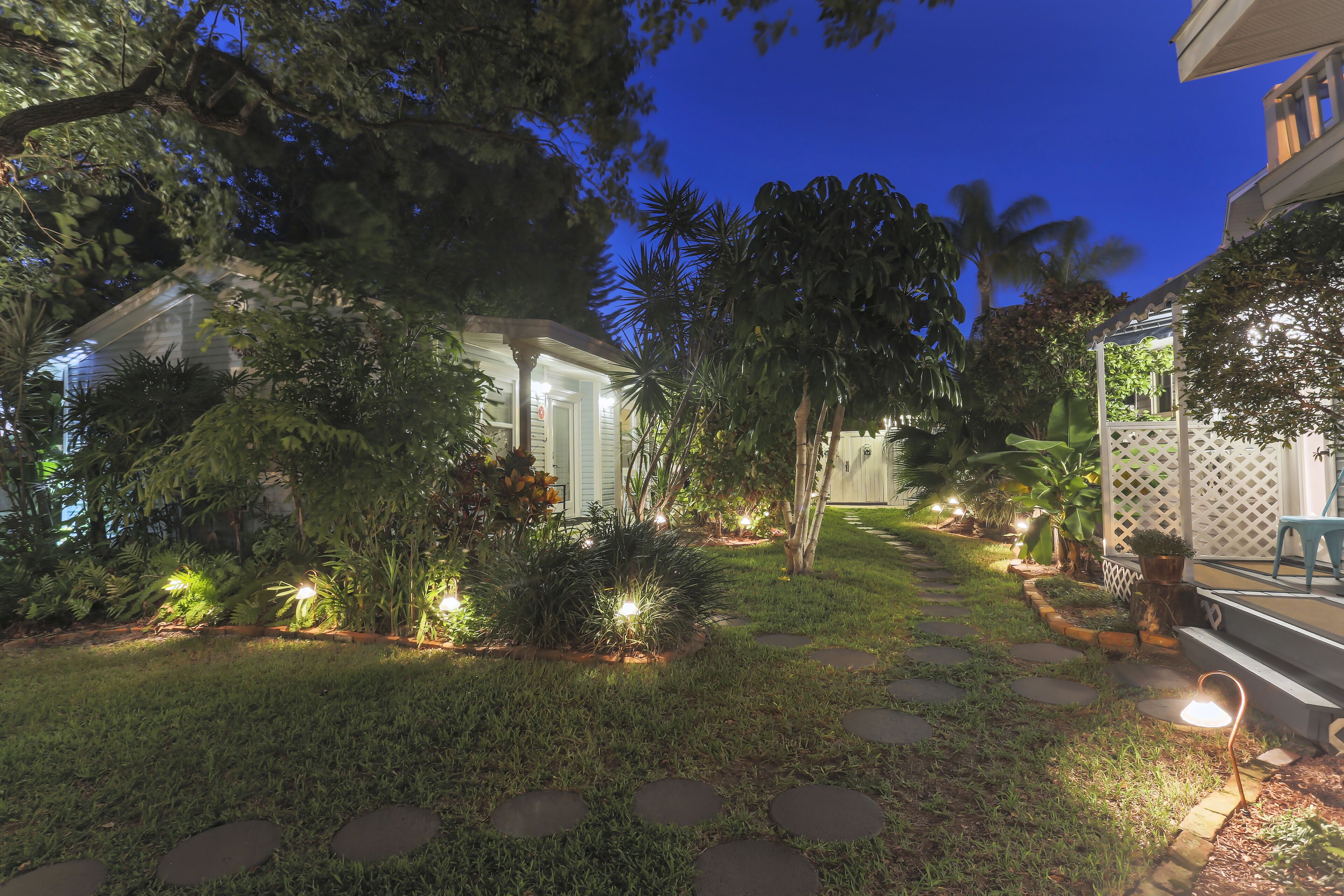 A well-lit garden pathway leading to a house with a porch, surrounded by lush greenery and palm trees.