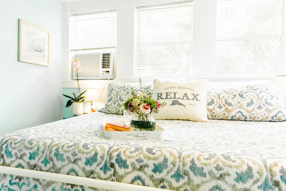 A neatly arranged bedroom with a bed, pillows, a bedside table, a lamp, and a window with blinds.