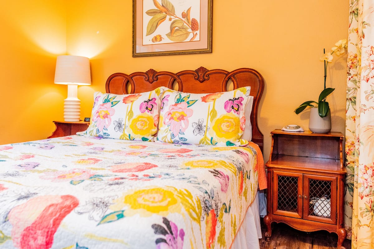 A bed with floral bedding, a bedside lamp, a framed botanical print on the wall, and a nightstand with a potted plant.