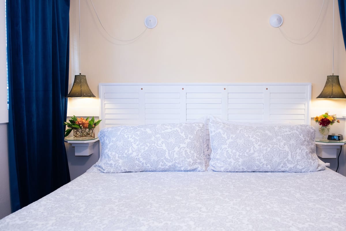 A neatly made bed with a patterned bedspread and pillows, flanked by two bedside tables with lamps and floral arrangements.