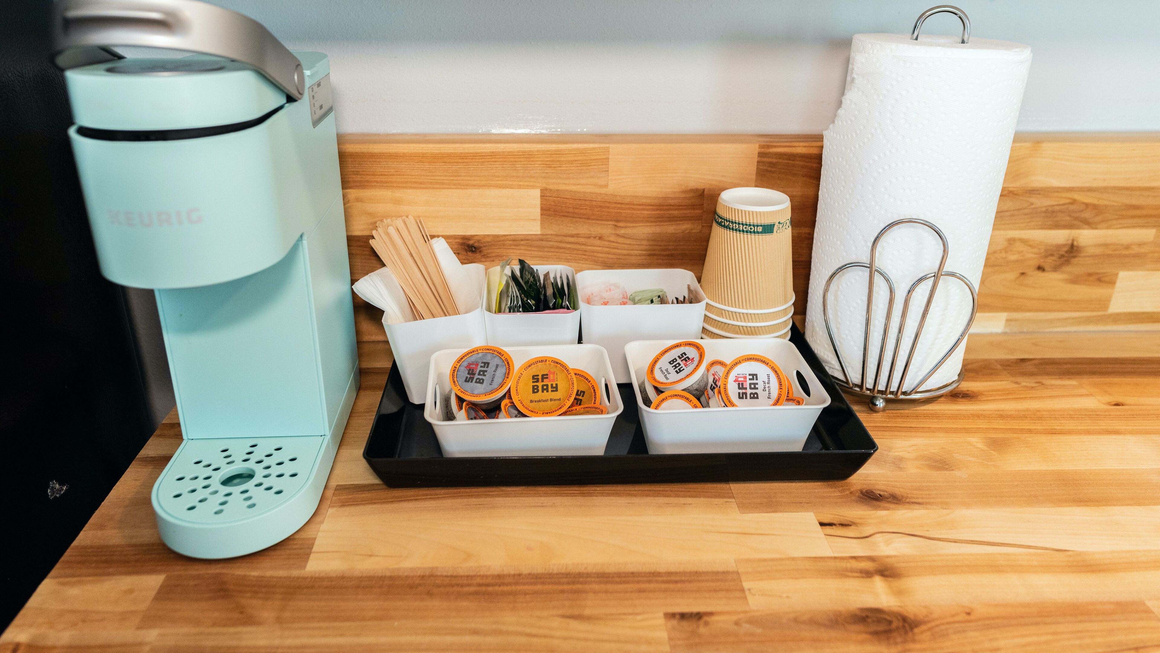 Una cafetera, una bandeja con tazas reutilizables y cubiertos, y un dispensador de toallas de papel sobre una encimera de madera.