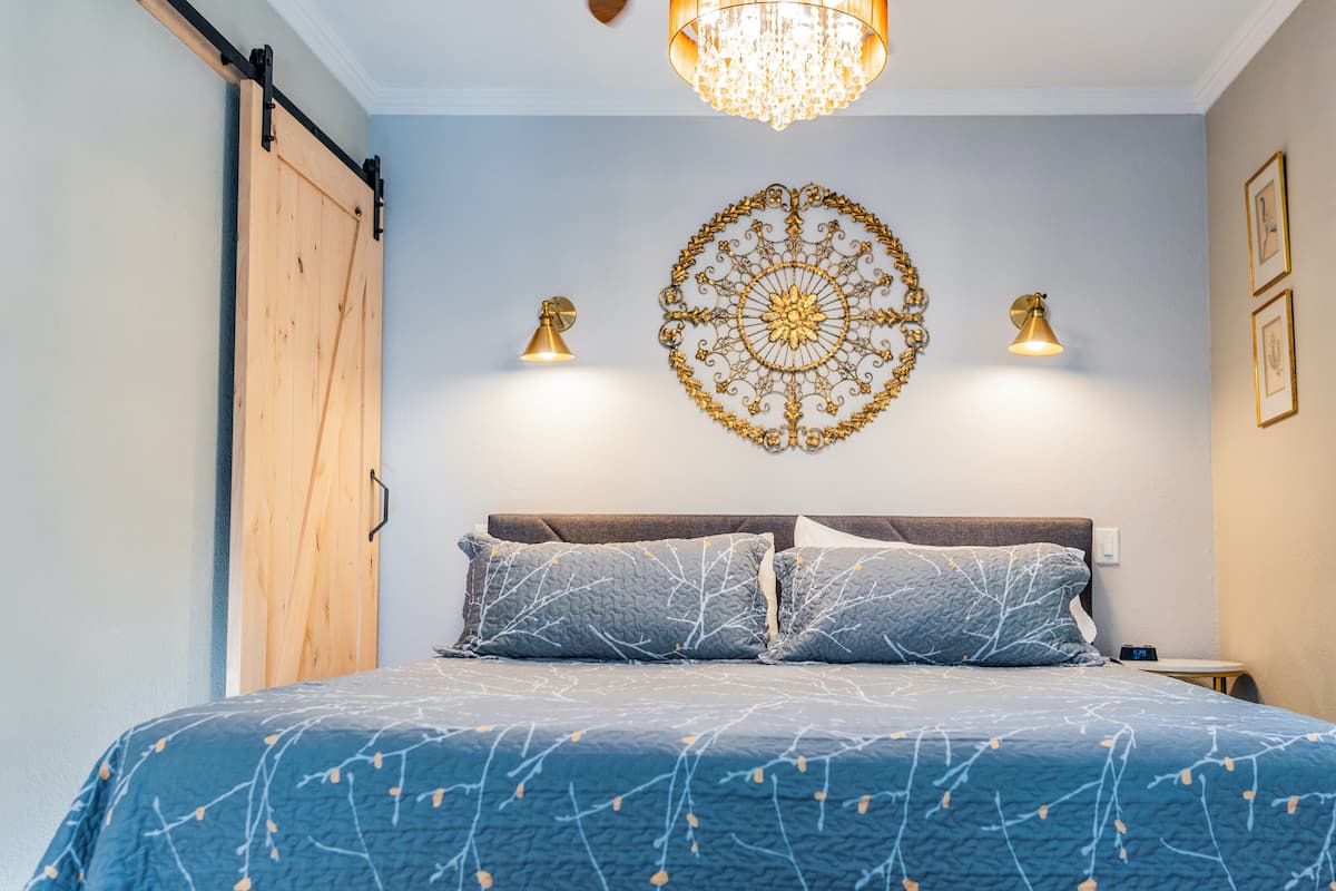 A bedroom with a bed, a chandelier, wall sconces, a wooden door, and a window with blinds.