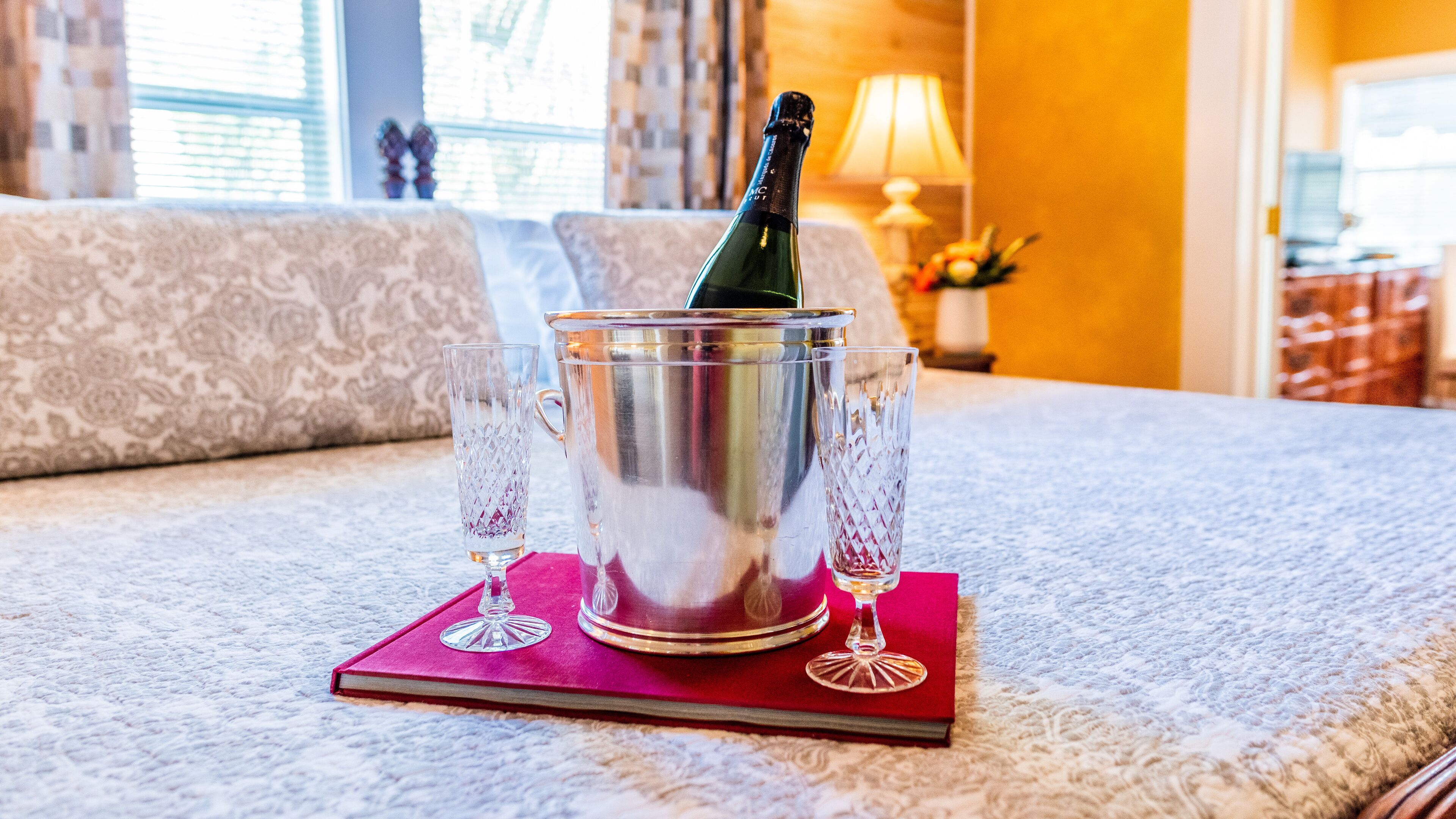 A champagne bottle in a metal bucket on a red coaster, surrounded by two crystal glasses, on a textured carpet in a room with patterned sofas and a lamp.