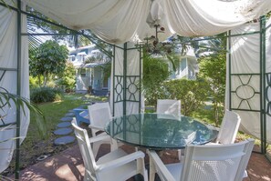 A glass-top table with white chairs in a covered outdoor area with a view of a house and greenery. - Meranova Guest Inn (Dunedin)