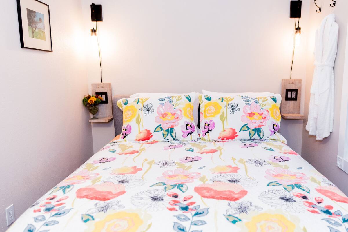 A bed with a floral bedspread, two wall-mounted lights, a framed picture, and a window with a view.