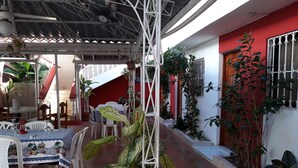 Un espace extérieur aménagé en terrasse, avec des tables et des chaises, des plantes en pot et un bâtiment rouge en arrière-plan.