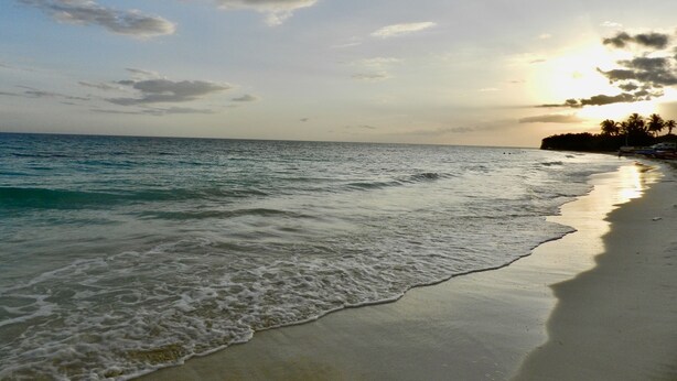 Une plage où les vagues viennent doucement lécher le rivage, un ciel dégagé et un coucher de soleil à l’horizon.