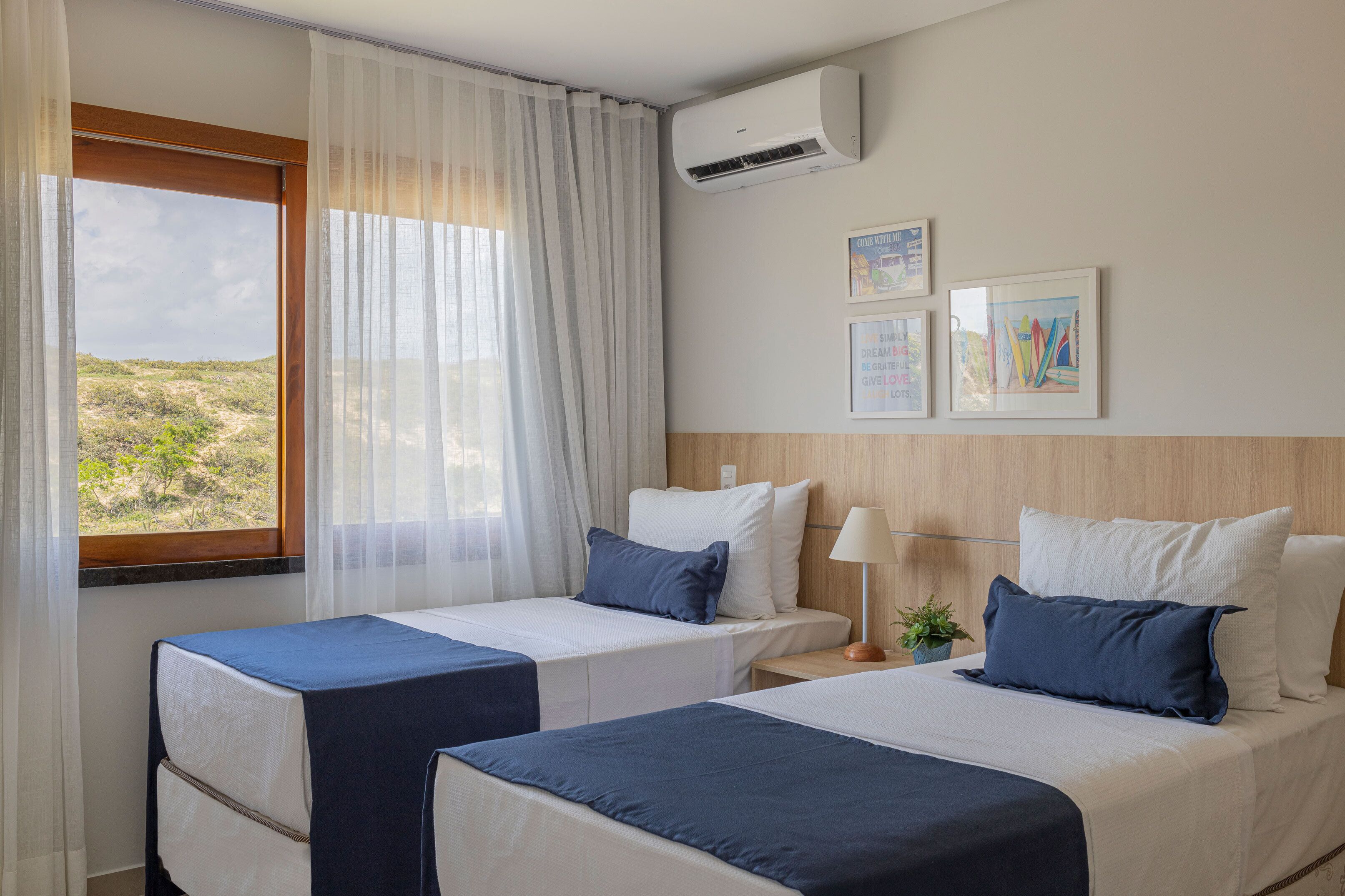 A hotel room with two beds, a window with sheer curtains, an air conditioning unit, and framed pictures on the wall.