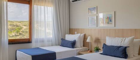 Quarto de hotel com duas camas, janela com cortinas translúcidas, aparelho de ar condicionado e quadros nas paredes.