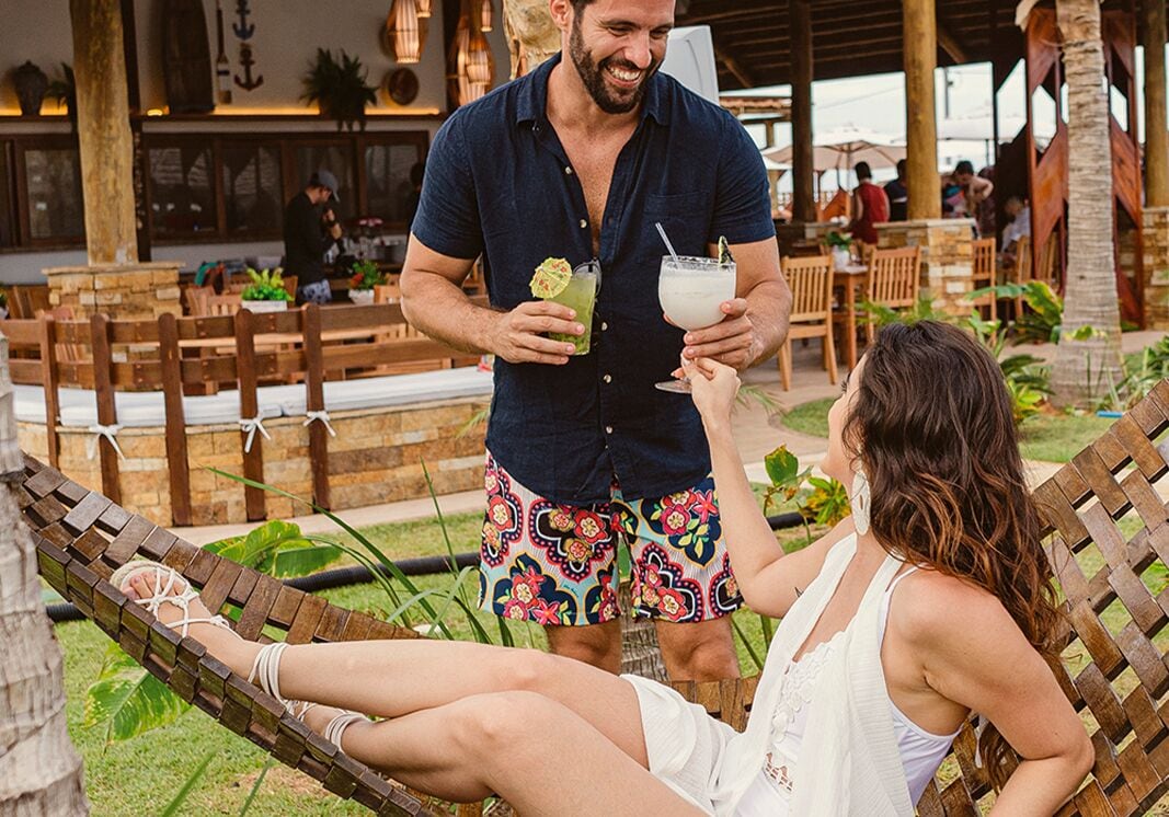 A couple relaxing on a hammock in an outdoor setting with wooden furniture and tropical plants.