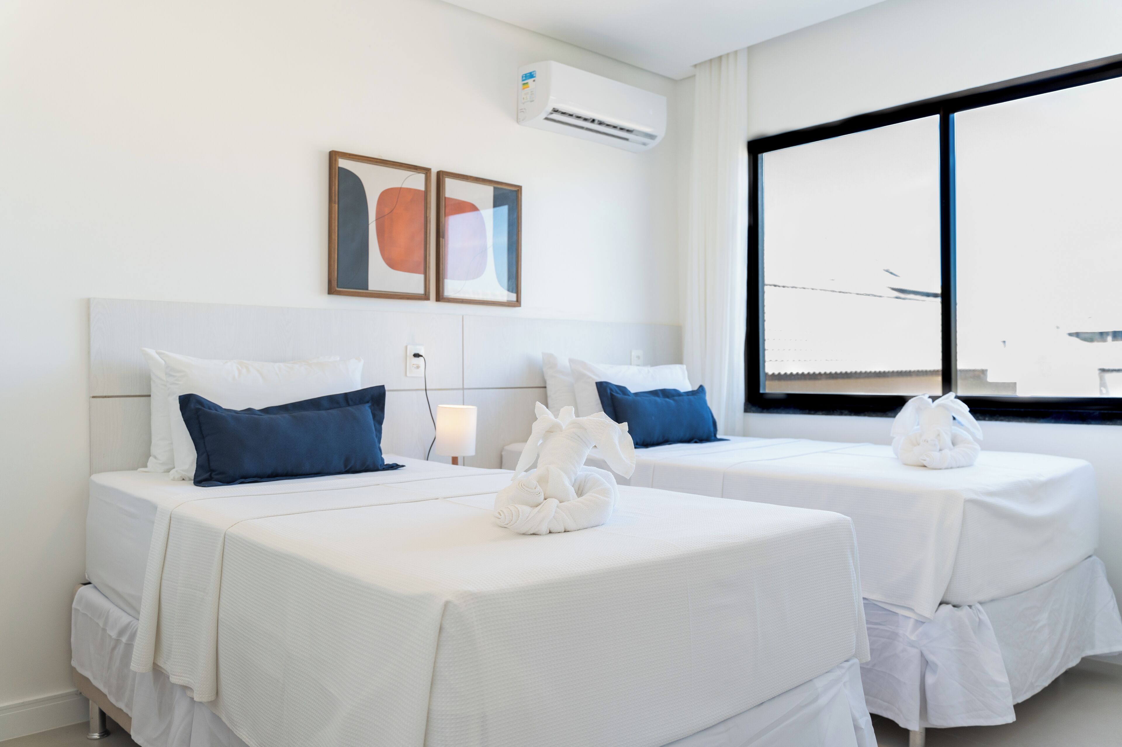 Habitación de hotel con dos camas, ropa de cama blanca, almohadas azules, una toalla blanca, aire acondicionado, una ventana con vista a un avión y dos cuadros abstractos en la pared.
