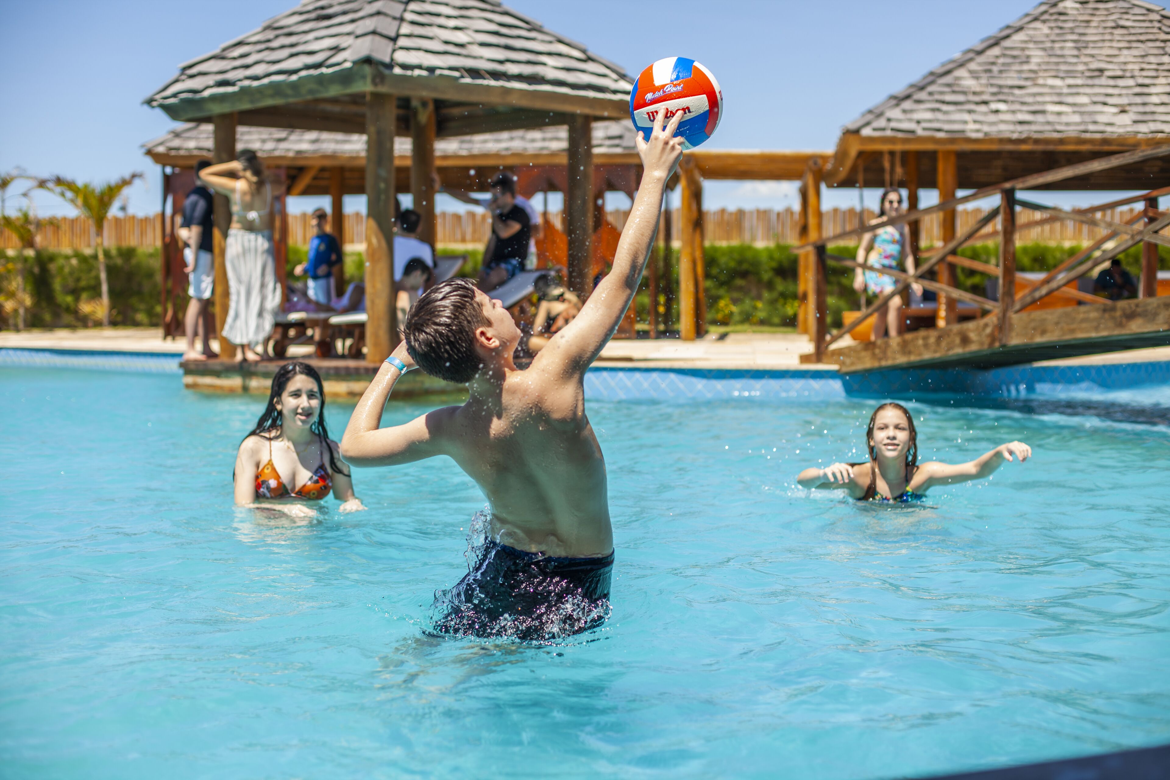 A pool area with a wooden structure, people in the water, and a volleyball.
