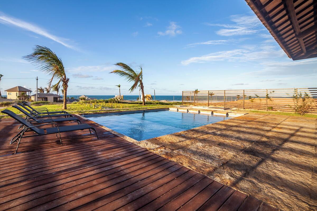 Un área de piscina con terraza de madera y sillones de descanso, rodeada de palmeras y vista al océano.