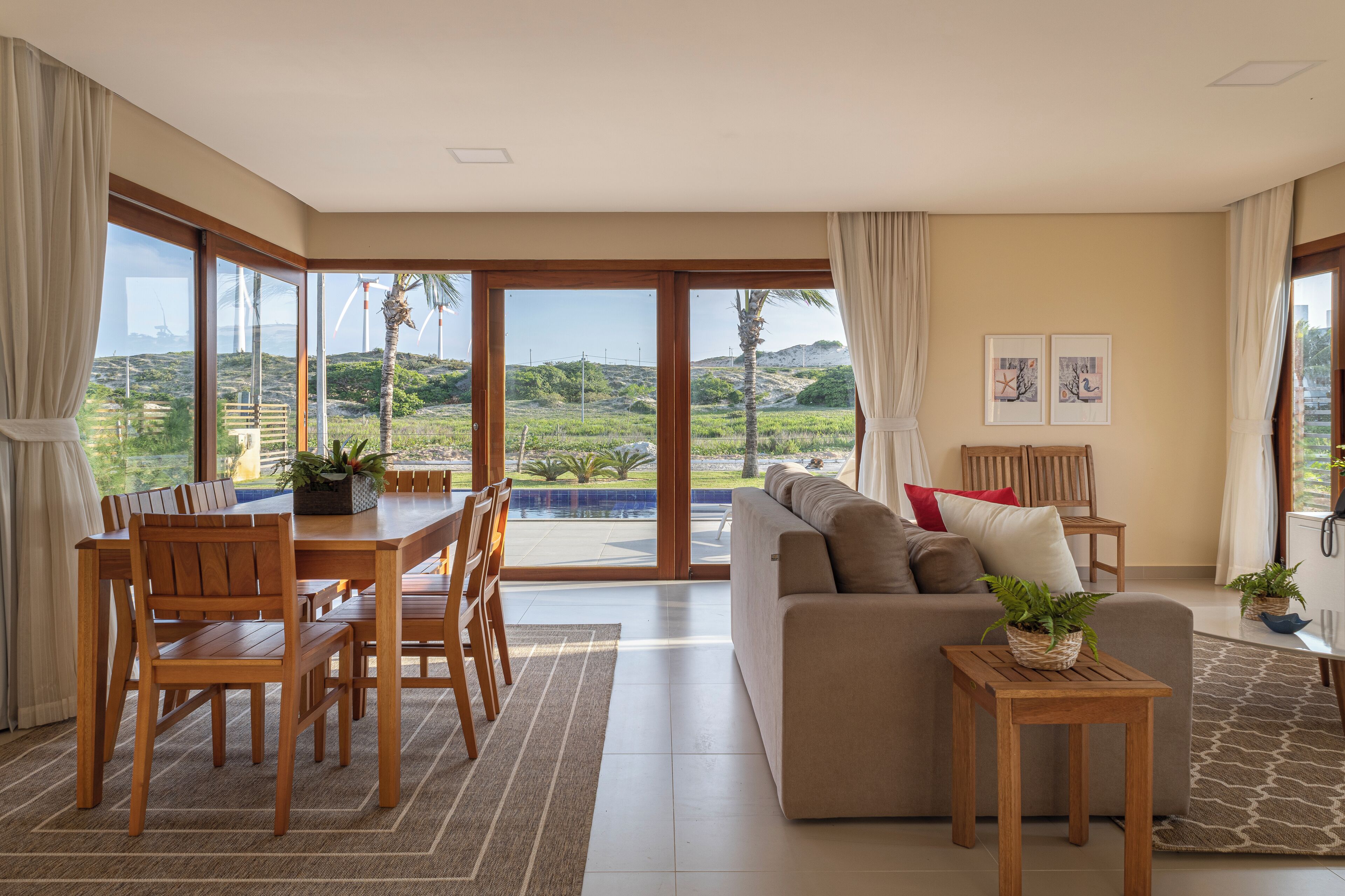 Amplia sala de estar con un gran ventanal corredera que da a una zona de piscina, muebles de madera y un sofá con cojines.