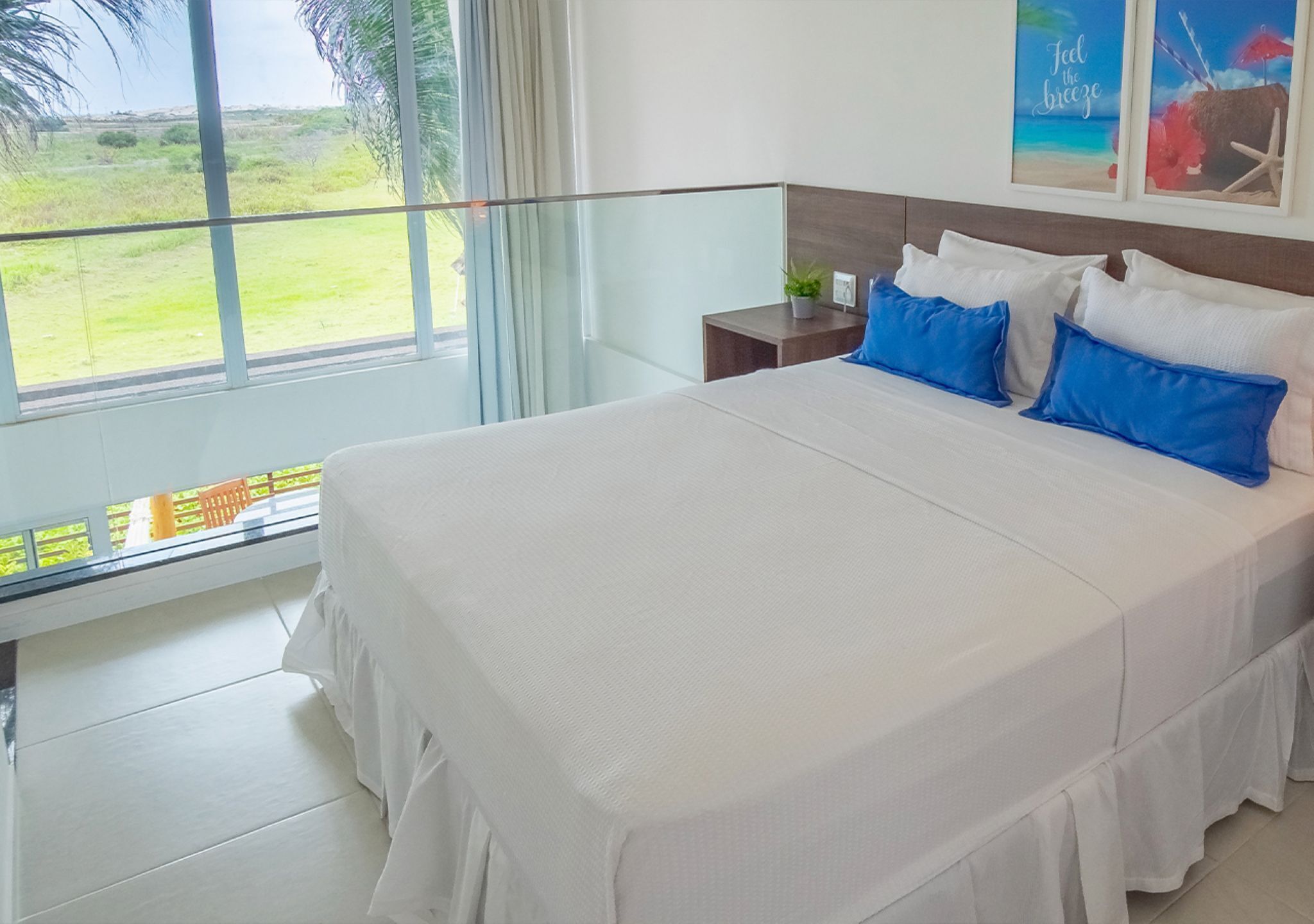 A bedroom with a large bed, a nightstand, a window with a view of a grassy area, and a wall with framed artwork.