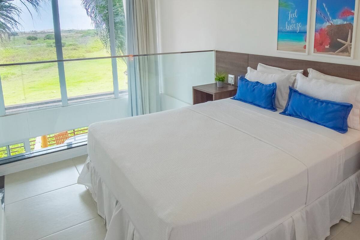 Un dormitorio con una cama grande, una mesita de noche, un ventanal con vistas a un espacio verde y una pared decorada con obras enmarcadas.