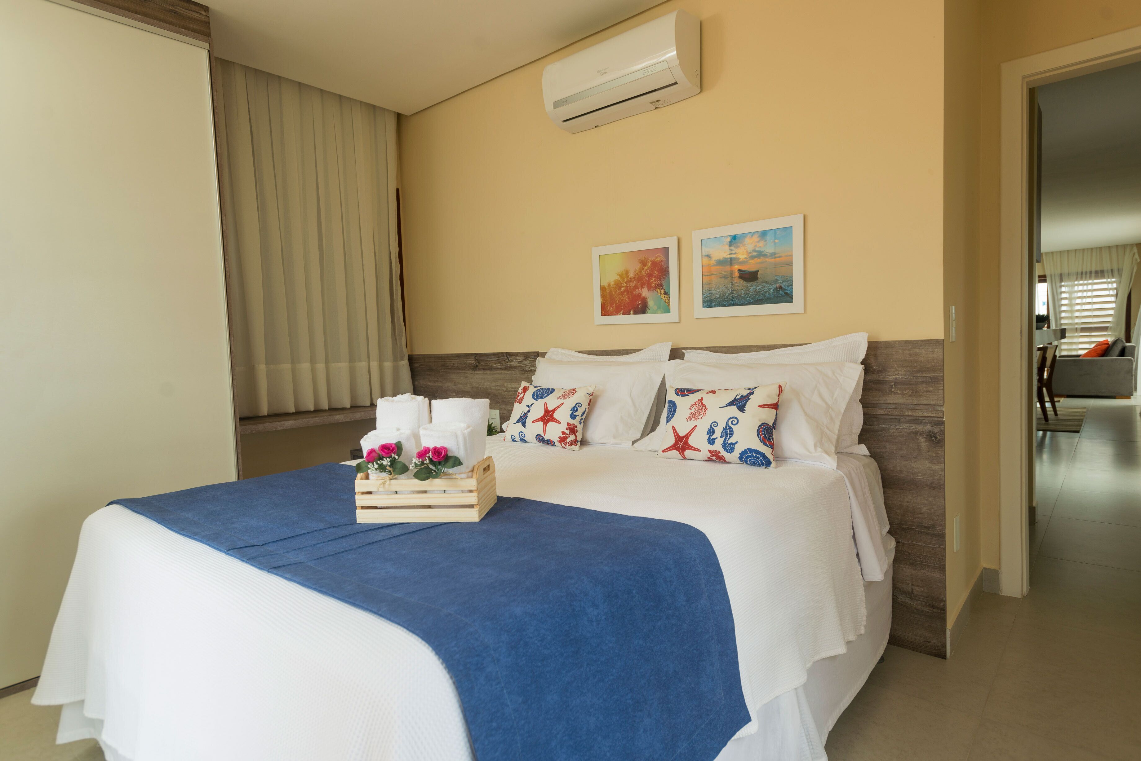 A hotel room with a bed, a wooden headboard, a blue bedspread, white bedding, two wall-mounted paintings, and a wooden side table with a tray of toiletries.