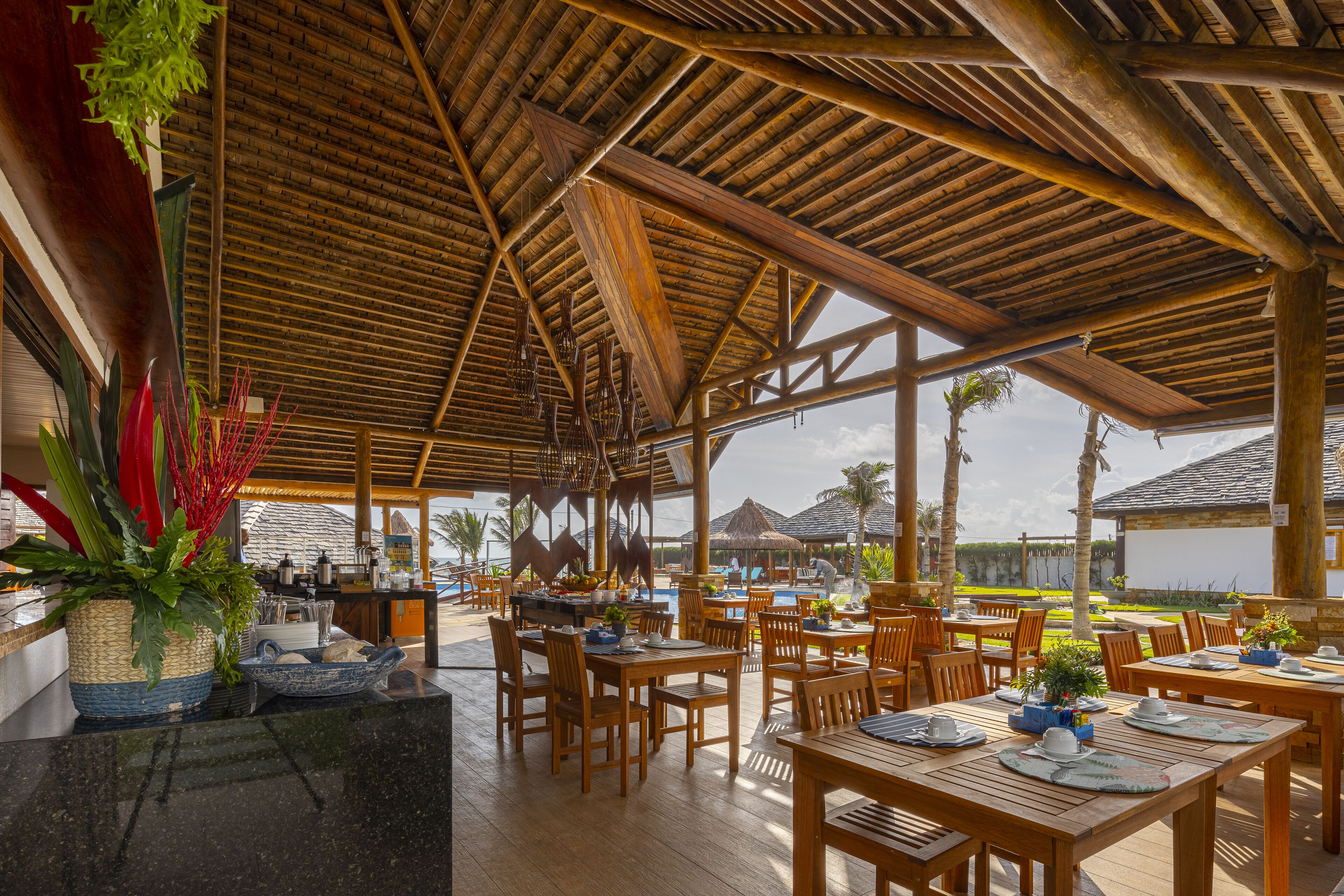 Un área de comedor al aire libre con mesas y sillas de madera, techo de paja y vista al océano.