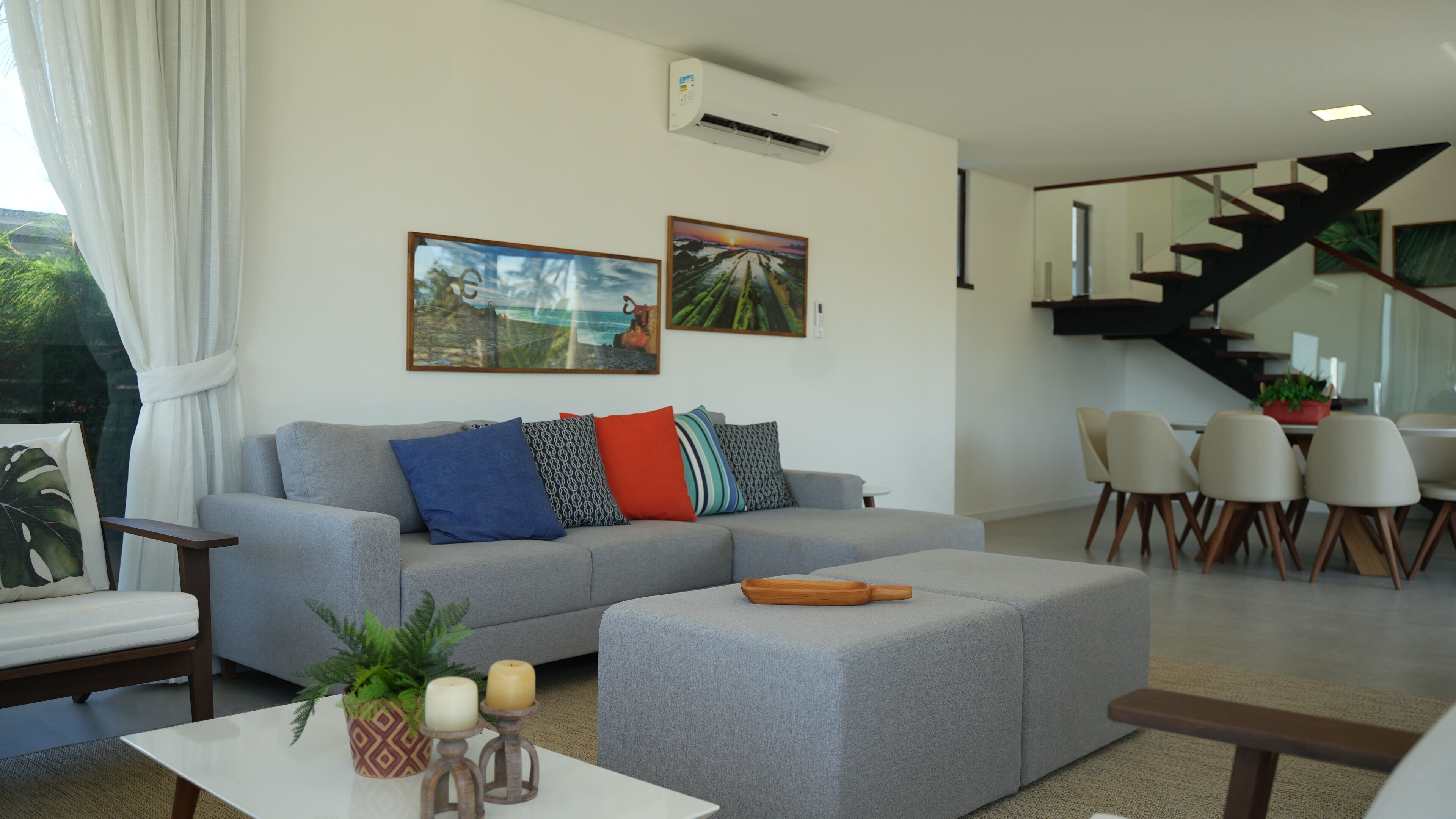A modern living room with a grey sofa, a coffee table, and a dining area with chairs. There are two paintings on the wall, a staircase, and an air conditioning unit.