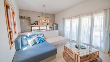Uma sala de estar moderna com sofá, mesa de centro de vidro e uma área de cozinha ao fundo.