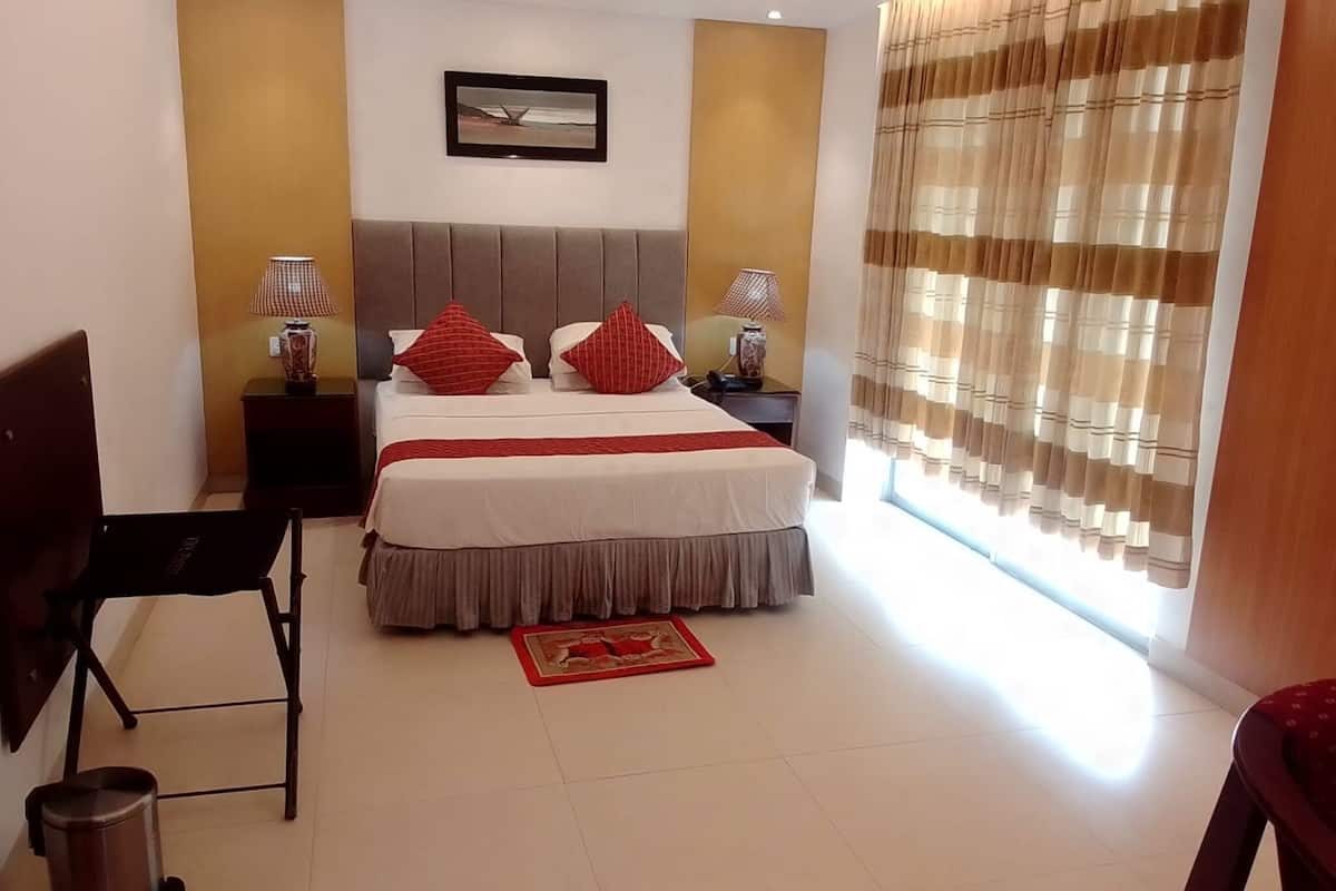 A hotel room with a bed, a desk, a chair, a trash bin, and a window with curtains.