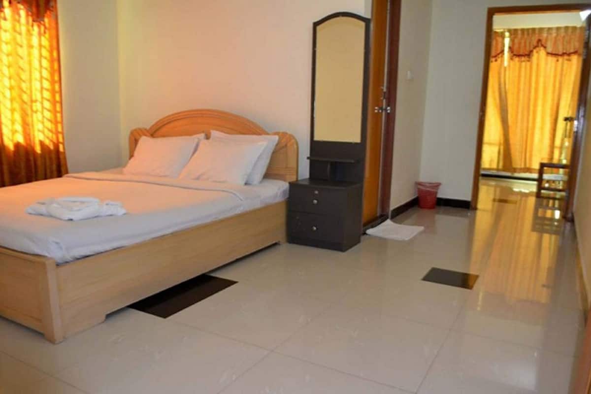 A neatly made bed with a wooden headboard, a bedside table with a mirror, and a red bucket on the floor.