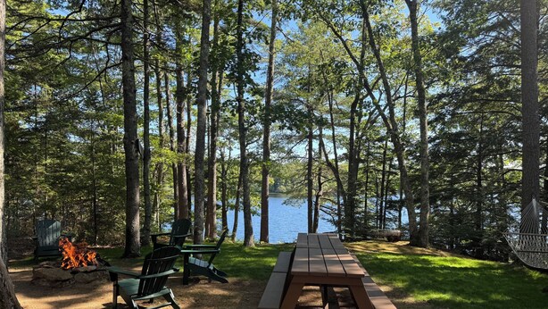 A picnic table with chairs and a fire pit in a forested area with a lake view.
