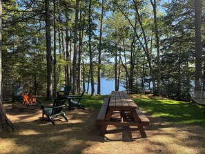A picnic table with chairs and a fire pit in a forested area with a lake view.