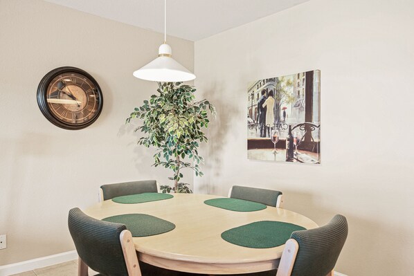 A dining area with a round table, four chairs, a wall clock, a hanging pendant light, a potted plant, and a framed picture.