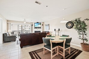 A living room with a brown leather sofa, a dining table with green chairs, a TV on a stand, and a potted plant.