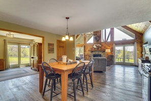 Un comedor amplio con mesa y sillas de madera, una cocina con chimenea de piedra y ventanas grandes que ofrecen vistas al exterior.