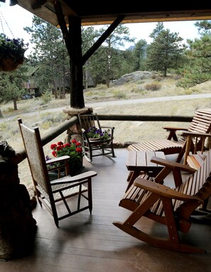 En træterrasse med gyngestole og et bord, med udsigt over et naturlandskab med træer og en vej.