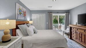 A bedroom with a bed, a television, a dresser, a chair, and a balcony with a view of greenery.