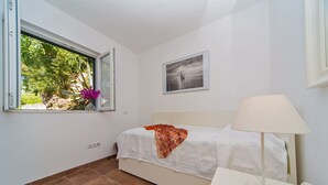 Ein Schlafzimmer mit Bett, Nachttisch, Lampe, einem Fenster mit Blick ins Grüne und einem gerahmten Bild an der Wand.