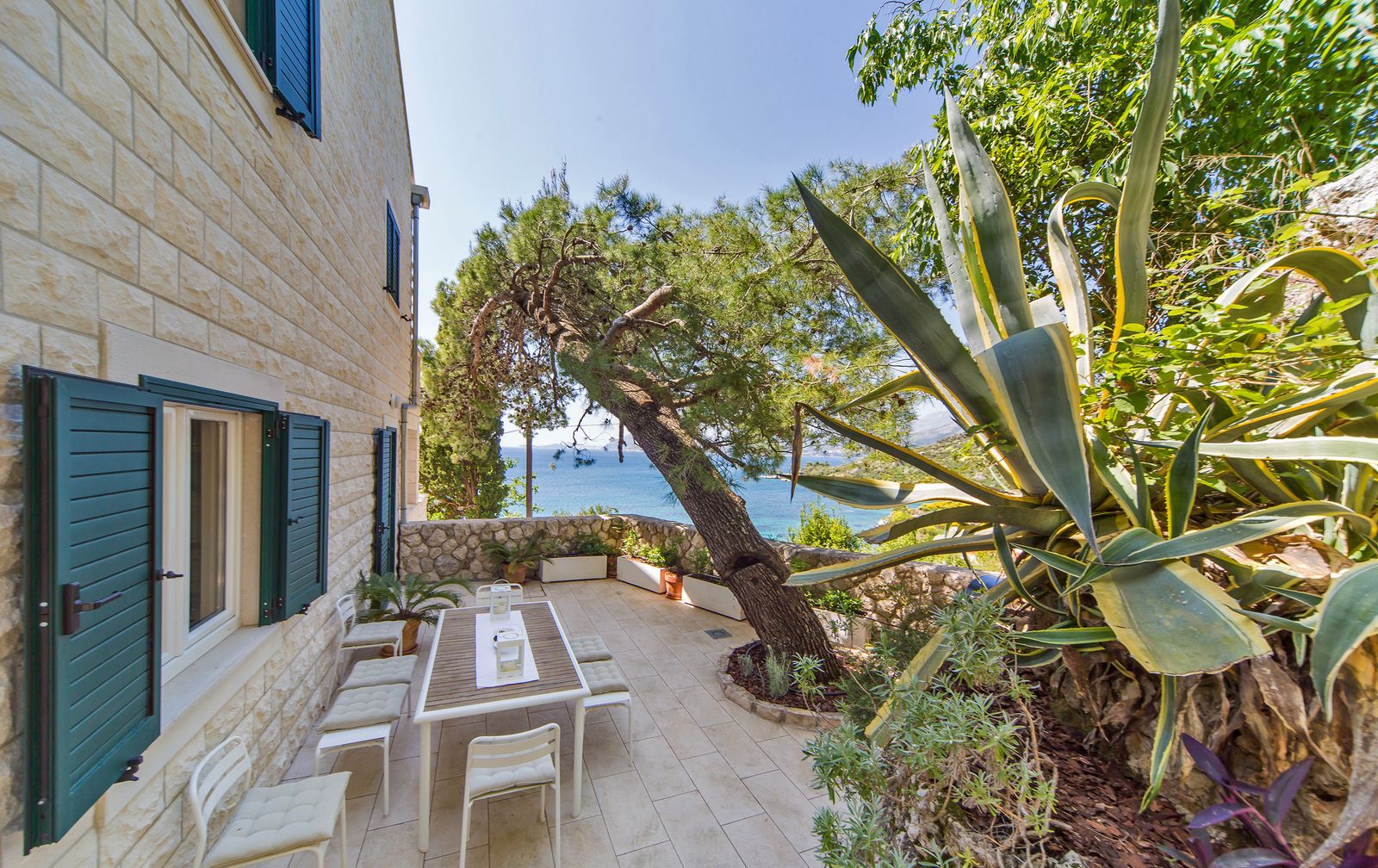 A stone building with a patio area featuring a table and chairs, surrounded by plants and overlooking a body of water.