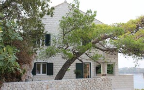A stone house with a sloped roof, green shutters, and a view of the water.