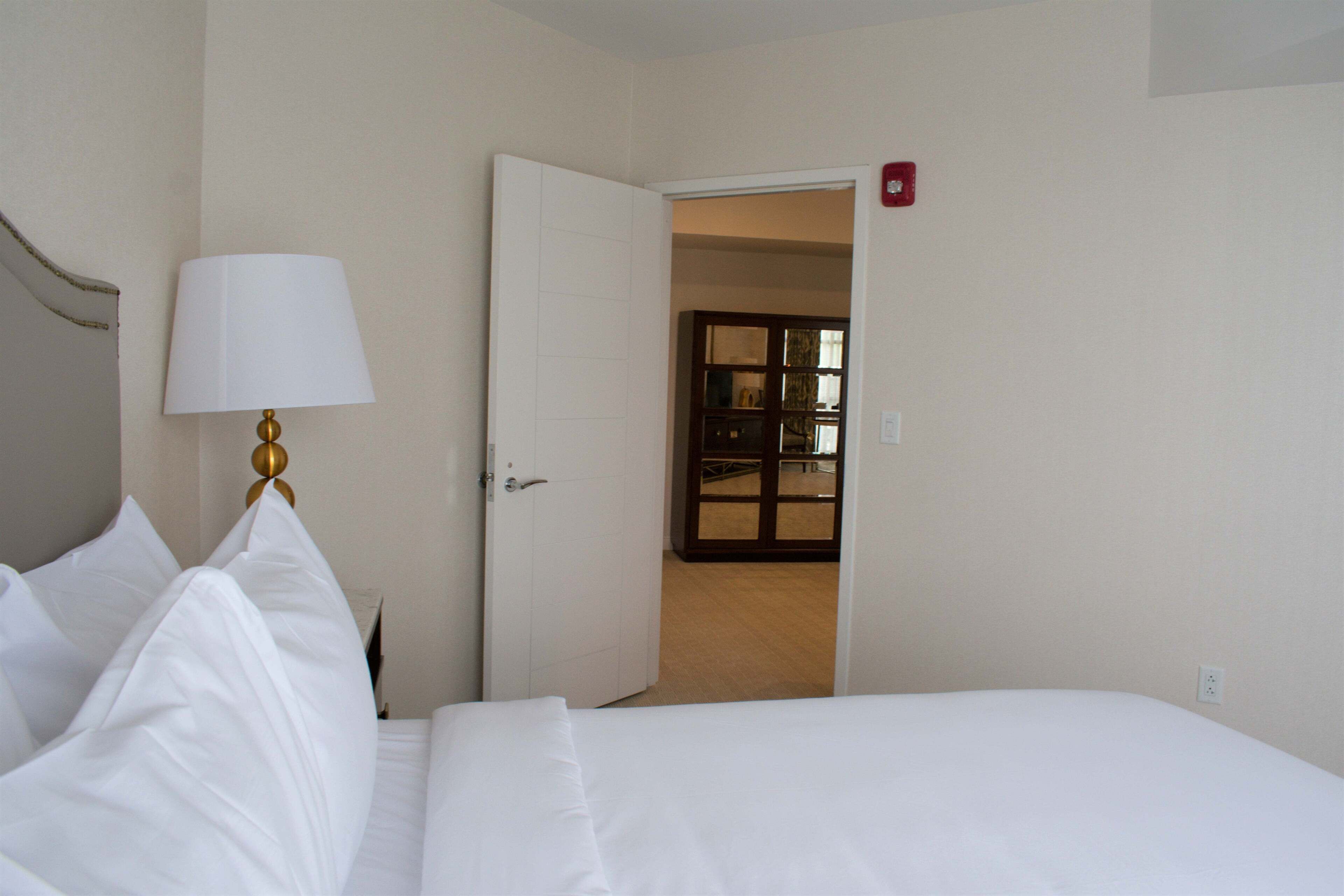 A hotel room with a bed, a lamp, and an open door leading to another room.
