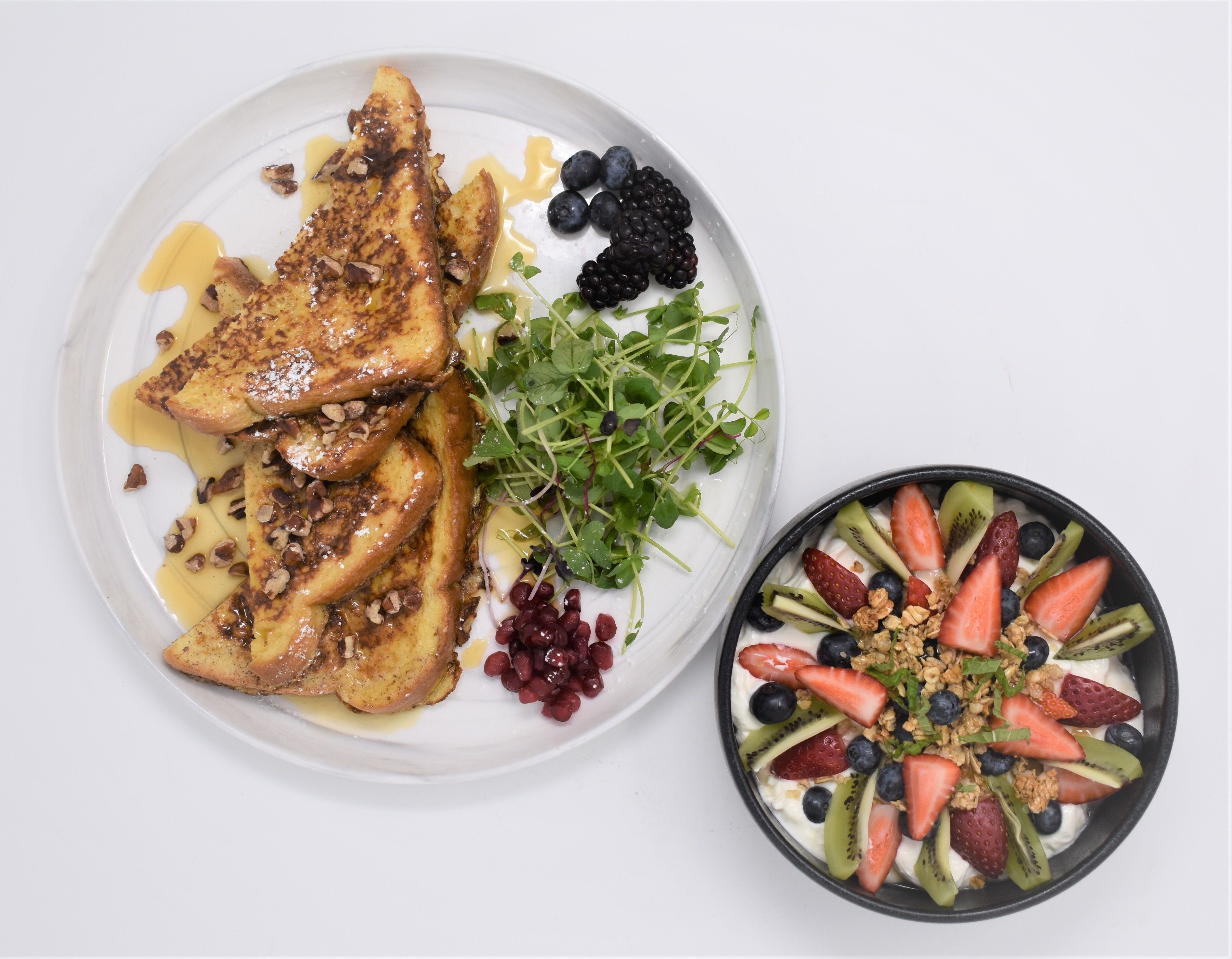 Una porción de tostadas francesas con almíbar y nueces, acompañada de un tazón de ensalada de frutas con fresas, arándanos y kiwi.