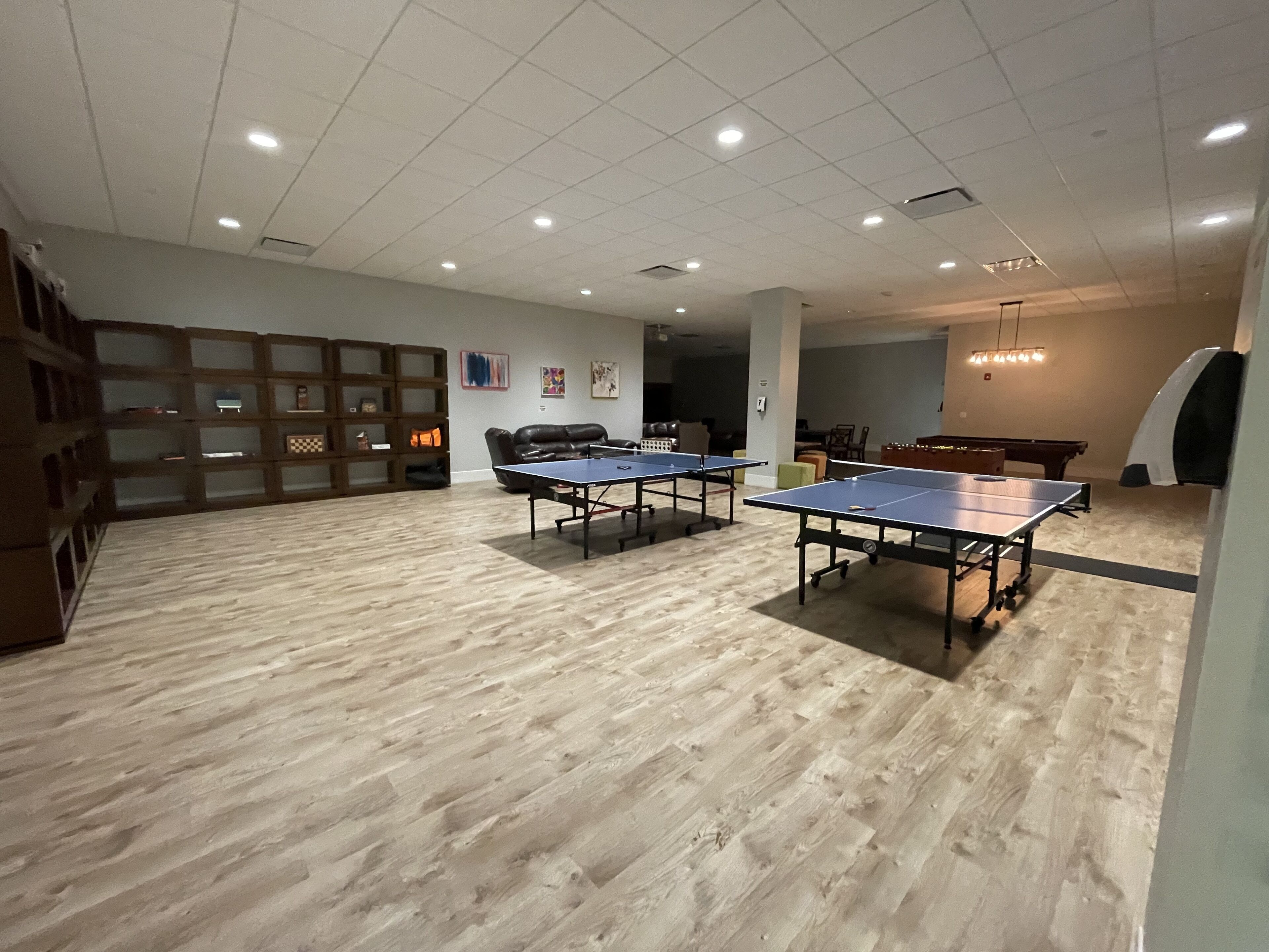 A spacious room with two ping pong tables, a billiards table, and a seating area with a sofa and chairs.