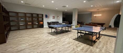 A spacious room with two ping pong tables, a billiards table, and a seating area with a sofa and chairs.