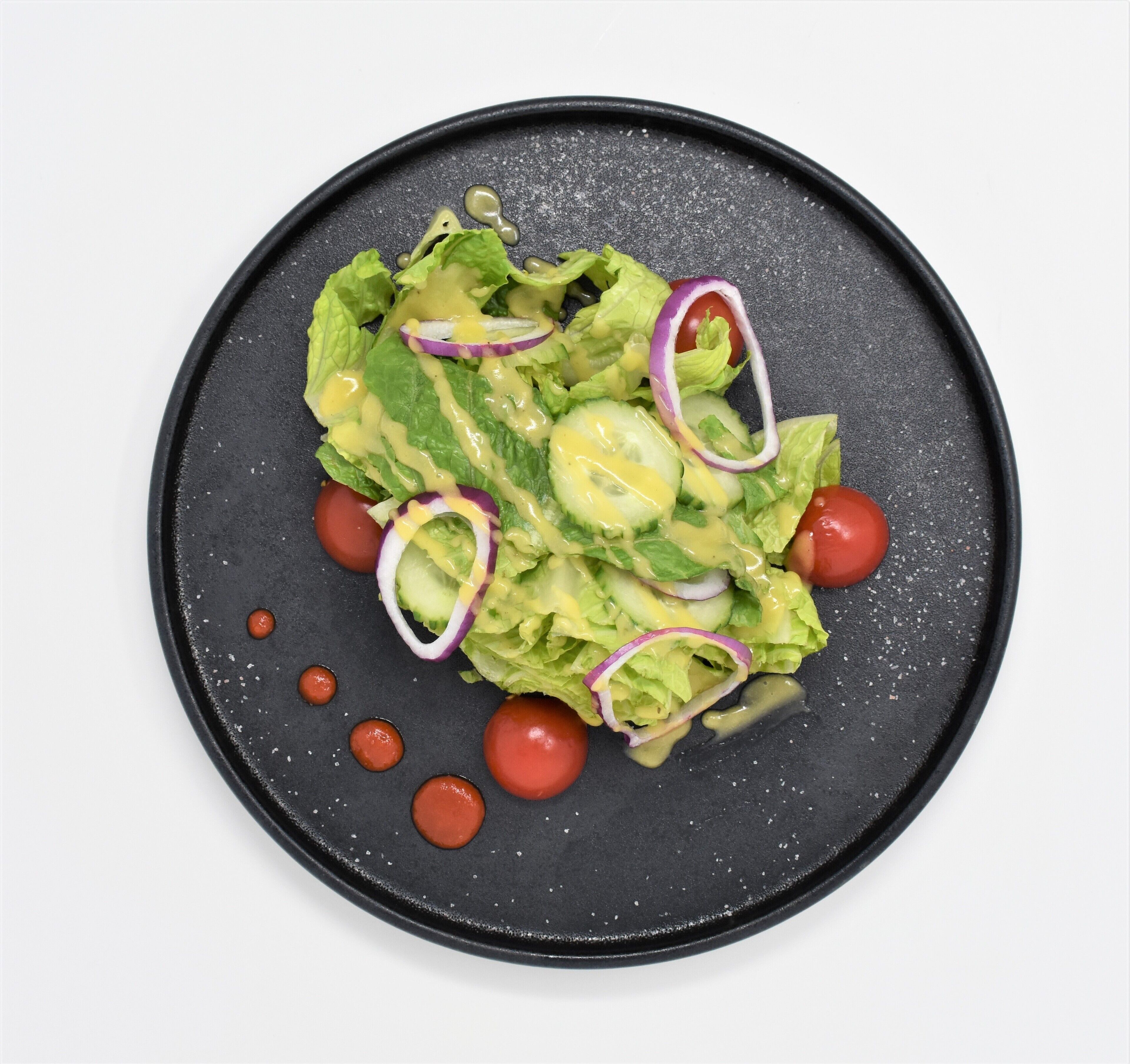 Un plato negro con una ensalada de lechuga, rodajas de pepino, aros de cebolla morada y tomates cherry, aderezada con una salsa cremosa.