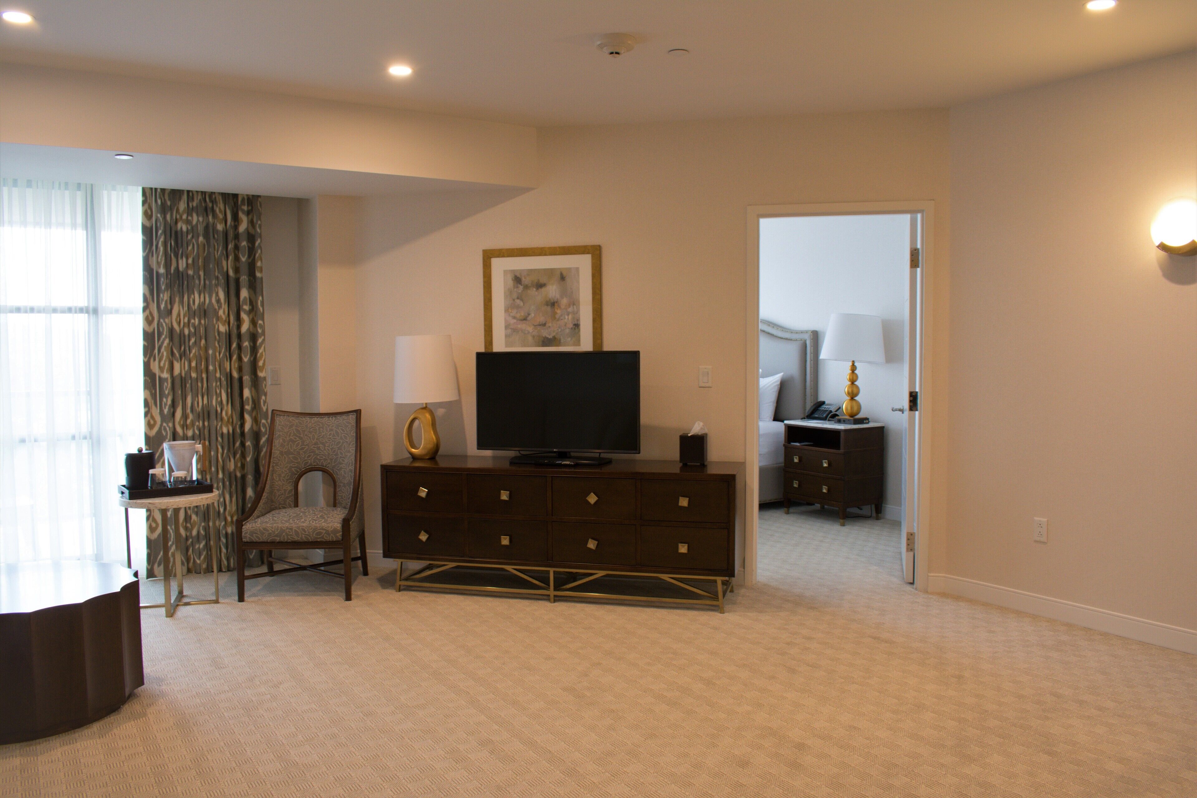 Habitación de hotel con televisor de pantalla plana, una silla, una mesita, un aparador y una puerta que da a otra habitación.