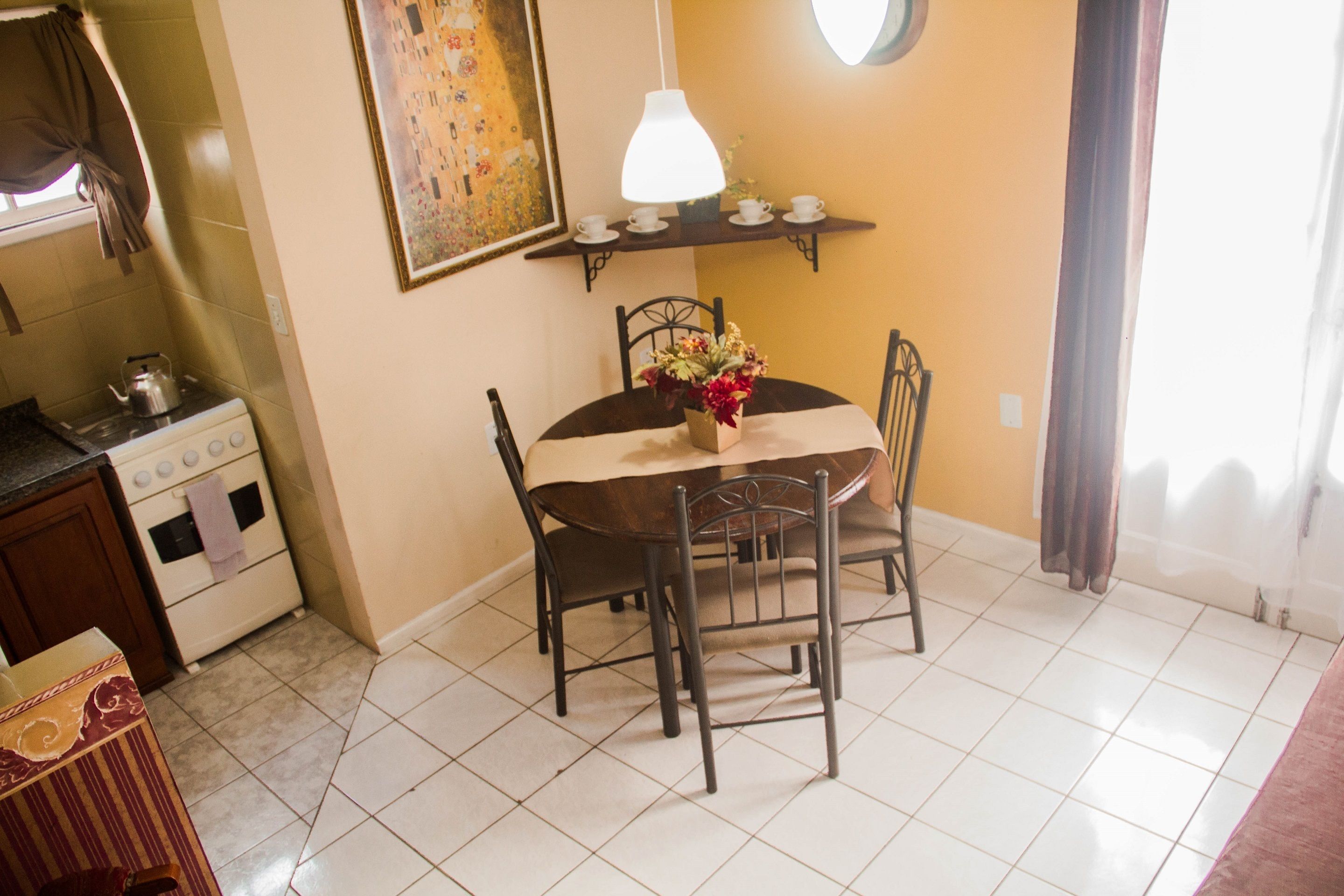A dining area with a round table, chairs, a vase with flowers, a hanging lamp, and a painting on the wall.