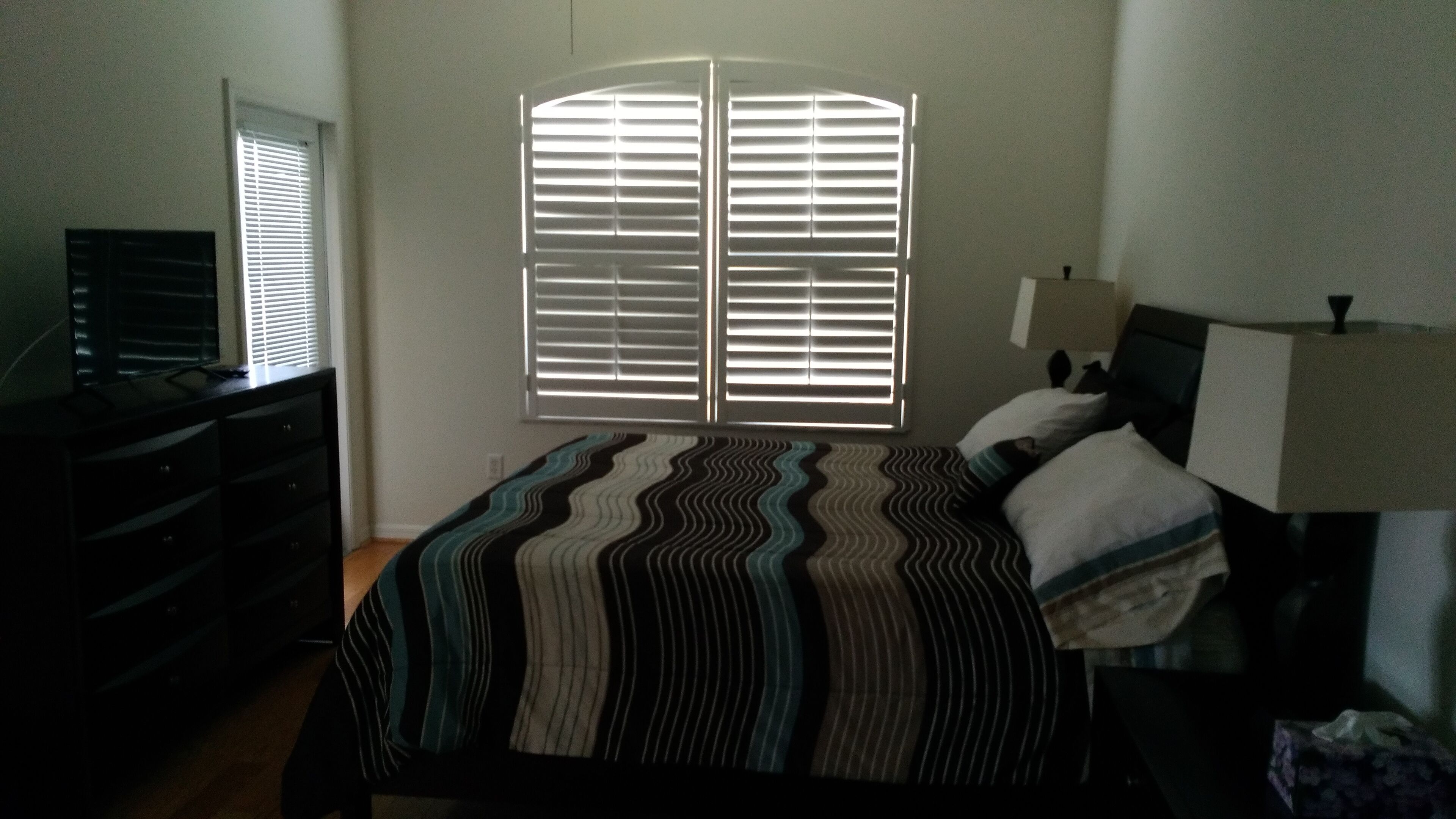 Ein Schlafzimmer mit Bett, Kommode, Rollläden und einer Lampe.