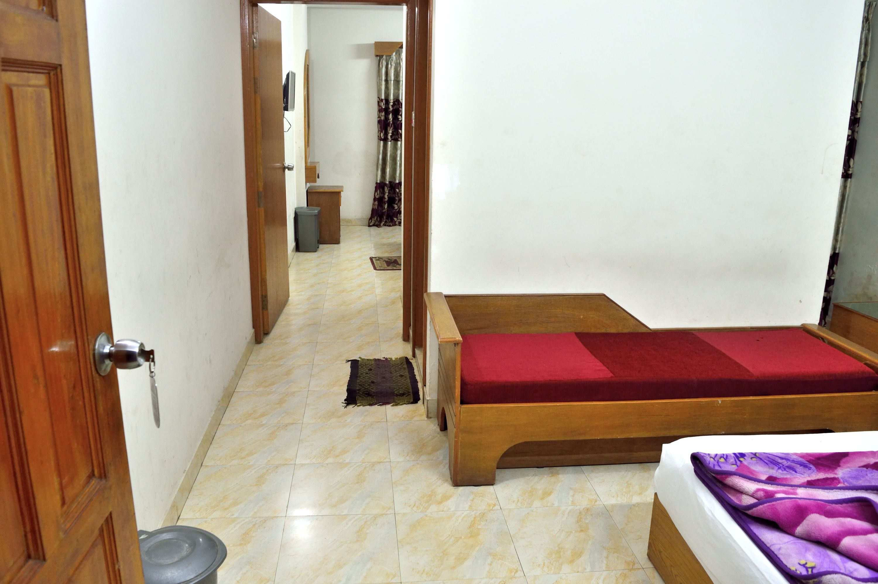 Una habitación con una cama de madera, colchón rojo, manta morada y suelo de baldosas.