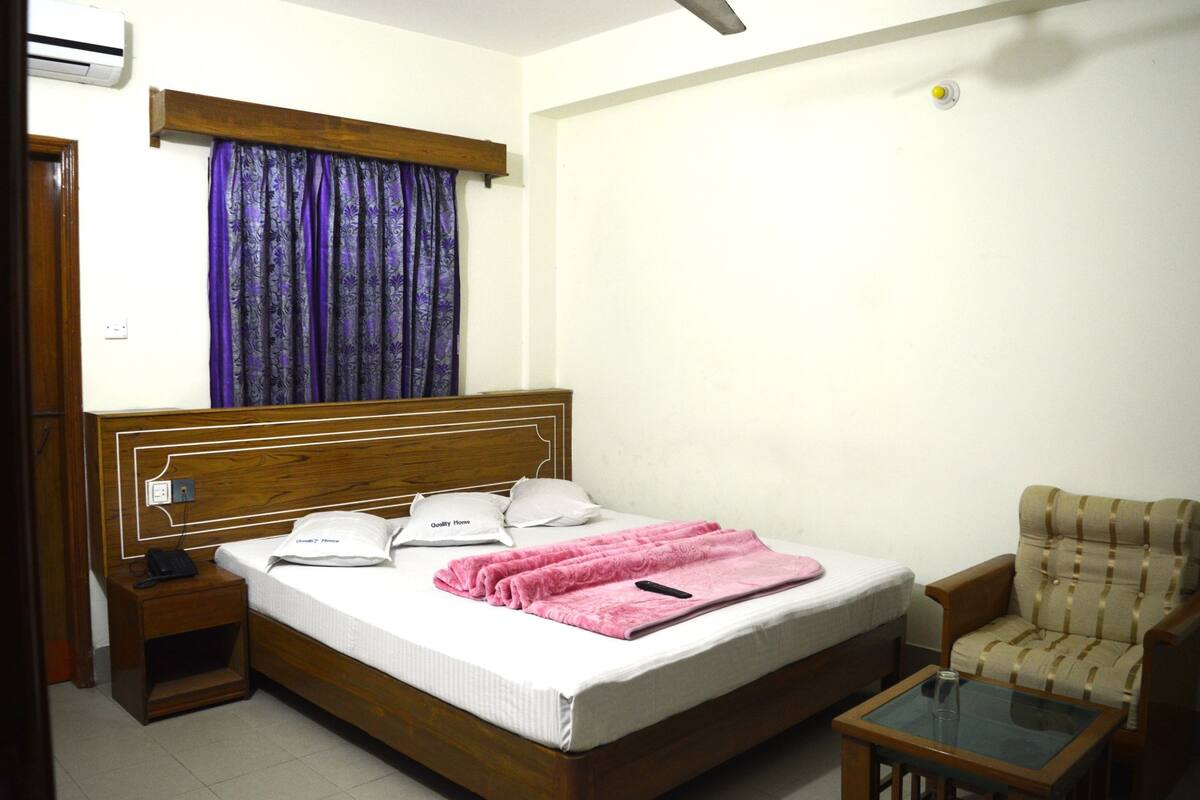 A neatly made bed with a pink blanket, a wooden headboard, a bedside table with a phone, and a chair with a glass table beside the bed.